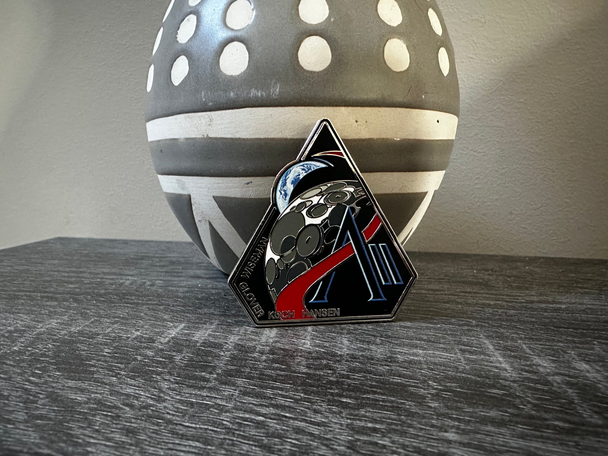 The Artemis II enamel pin, which depicts the Moon and the Earth, along with the letter A and II. Along the bottom edge of the pin are the last names of the 4 astronauts who will be onboard, Wiseman, Glover, Koch, Hansen. The pin is being propped up by a ceramic vase that is grey with white lines and white circles. It sits atop a gray wooden table.