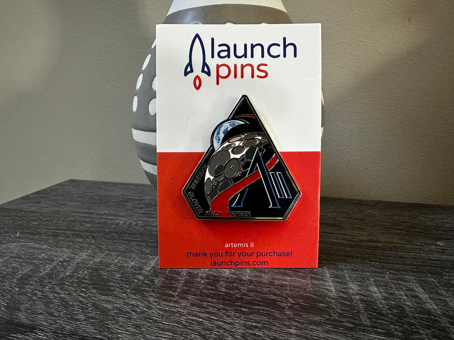 The Artemis II enamel pin, which depicts the Moon and the Earth, along with the letter A and II. Along the bottom edge of the pin are the last names of the 4 astronauts who will be onboard, Wiseman, Glover, Koch, Hansen. The pin is attached to a backing card. The top part of the backing card is white, with the name Launch Pins. The bottom half of the card is orange. The pin is being propped up by a ceramic vase that is grey with white lines and white circles. It sits atop a gray wooden table.