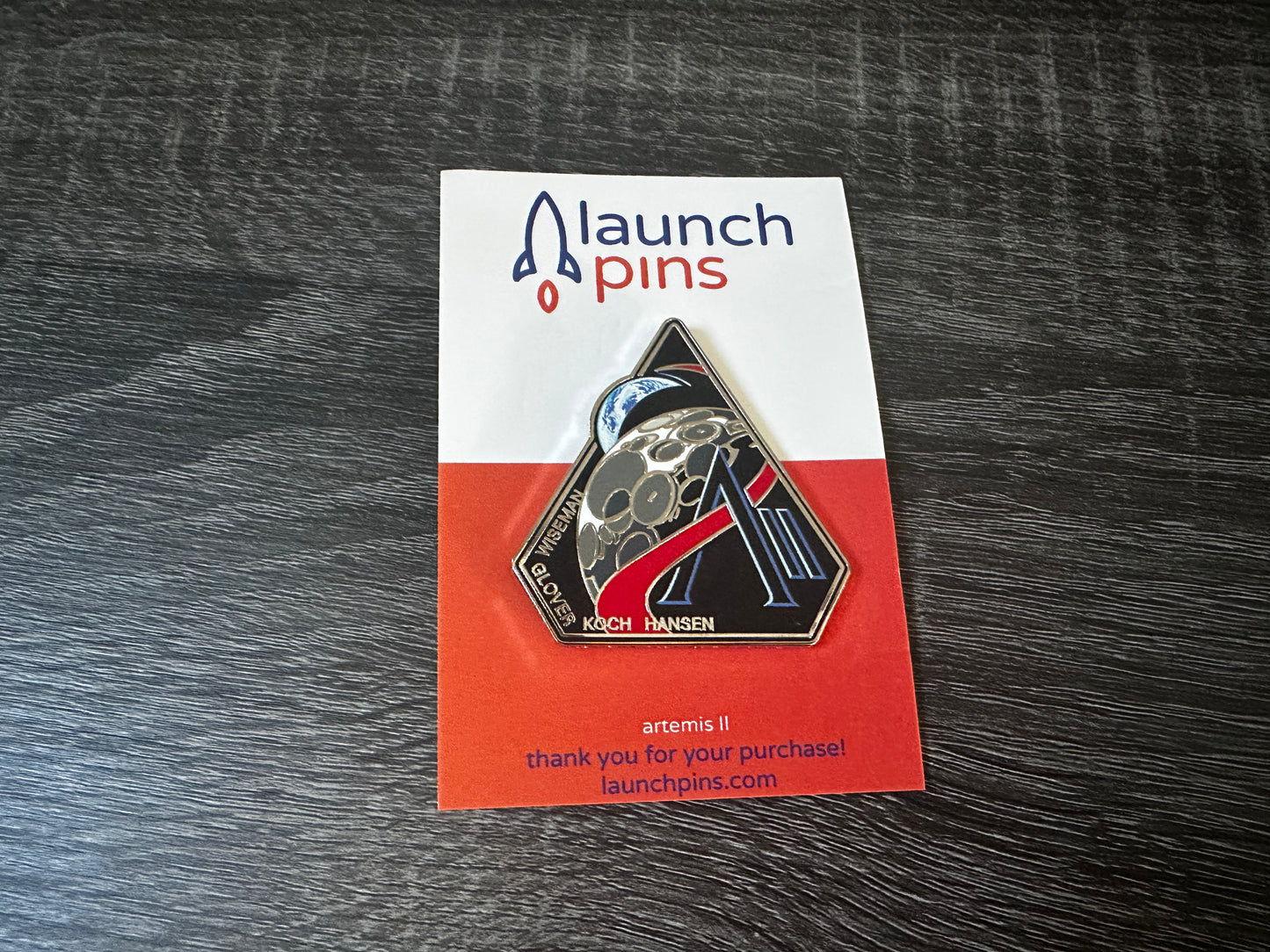 The Artemis II enamel pin, which depicts the Moon and the Earth, along with the letter A and II. Along the bottom edge of the pin are the last names of the 4 astronauts who will be onboard, Wiseman, Glover, Koch, Hansen. The pin is attached to a backing card. The top part of the backing card is white, with the name Launch Pins. The bottom half of the card is orange. The pin is lying atop a gray wooden table.