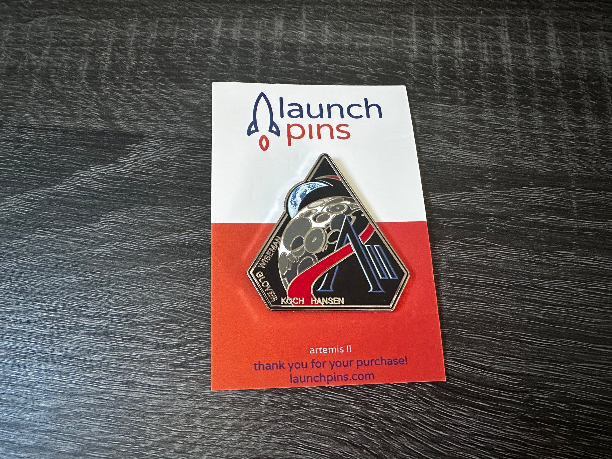 The Artemis II enamel pin, which depicts the Moon and the Earth, along with the letter A and II. Along the bottom edge of the pin are the last names of the 4 astronauts who will be onboard, Wiseman, Glover, Koch, Hansen. The pin is attached to a backing card. The top part of the backing card is white, with the name Launch Pins. The bottom half of the card is orange. The pin is lying atop a gray wooden table.